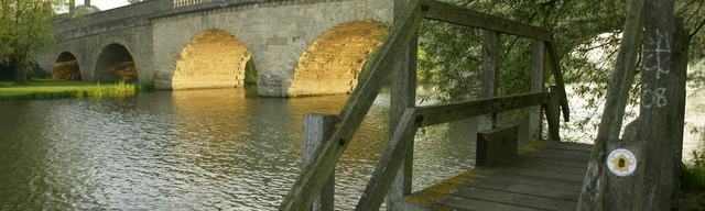 A view of a rural stone bridge with many arches, and a riverside footpath in the foreground