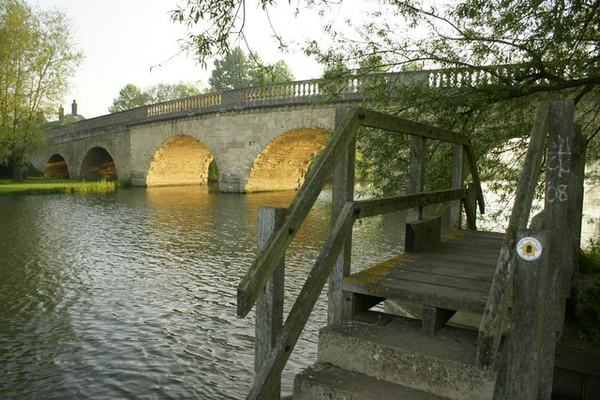 A view of a rural stone bridge with many arches, and a riverside footpath in the foreground