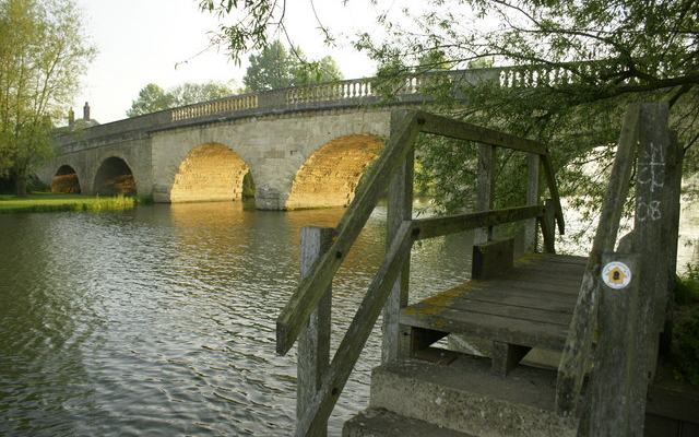 A view of a rural stone bridge with many arches, and a riverside footpath in the foreground